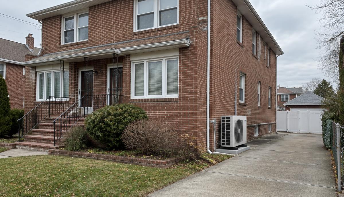 Air-source heat pump outdoor unit beside a brick two-family house in Queens